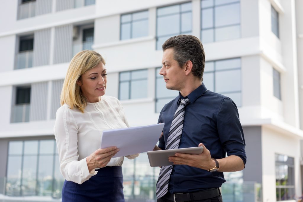 Content business partners discussing documents outdoors. Businesspeople standing, holding tablet computer and papers with building in background. Business partners concept.